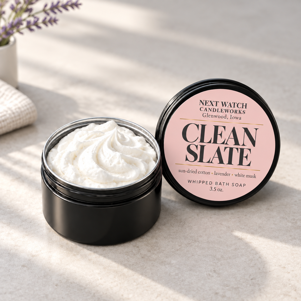 An open black jar of whipped bath soap showing pink soap inside, next to its lid which features a pink label reading 'NEXT WATCH CANDLEWORKS CLEAN SLATE'. The scene is styled on a textured beige surface with soft shadows and a hint of lavender in the background.