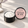 An open black jar of whipped bath soap showing pink soap inside, next to its lid which features a pink label reading 'NEXT WATCH CANDLEWORKS CLEAN SLATE'. The scene is styled on a textured beige surface with soft shadows and a hint of lavender in the background.