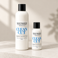 Two white bottles of Next Watch Candleworks Clean Cut For Him face and body lotion, one 8 oz and one 2 oz, standing on a light marble surface with soft shadow overlays.