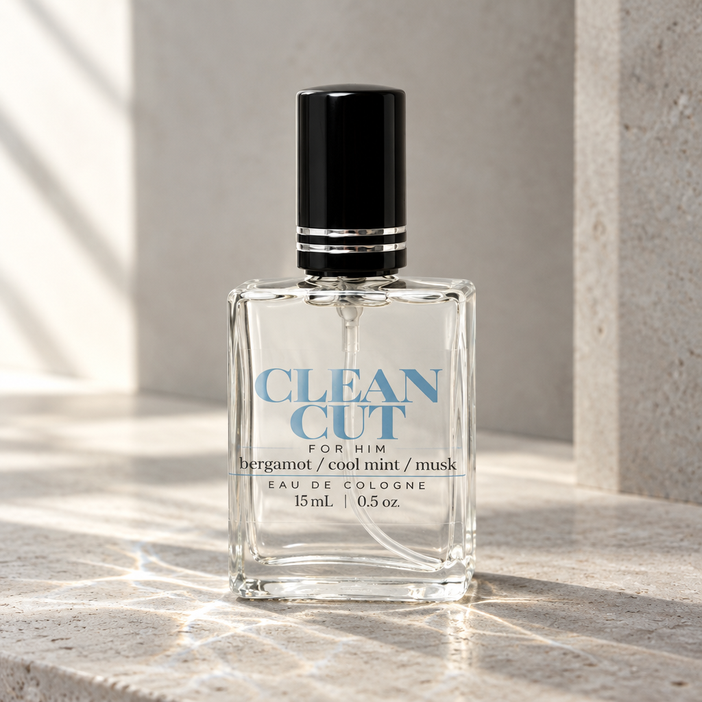 A 15ml bottle of Clean Cut Eau de Cologne for him, featuring a clear glass body, blue lettering, and a black cap, set on a textured stone surface with dramatic diagonal sunlight.