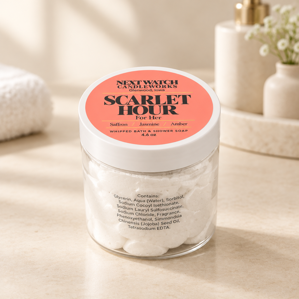 A clear glass jar of Next Watch Candleworks 'Scarlet Hour' whipped bath and shower soap, filled with white lather, set on a light beige marble surface with a soft-focus bathroom background.