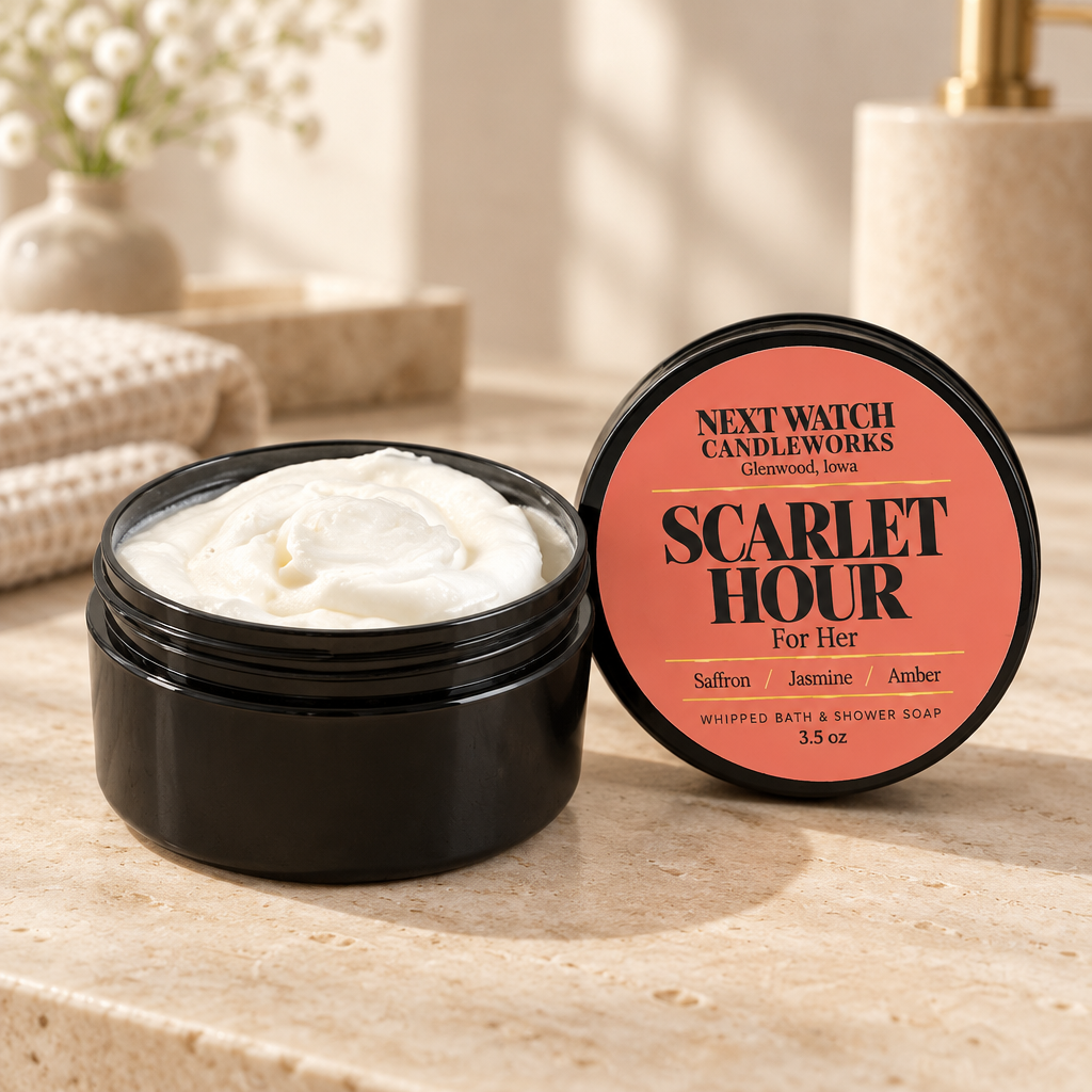 An open black jar of whipped bath and shower soap showing the white cream inside, next to its lid which features a coral-pink label reading 'NEXT WATCH CANDLEWORKS SCARLET HOUR For Her'. The scene is set on a beige stone countertop with a softly blurred bathroom background.