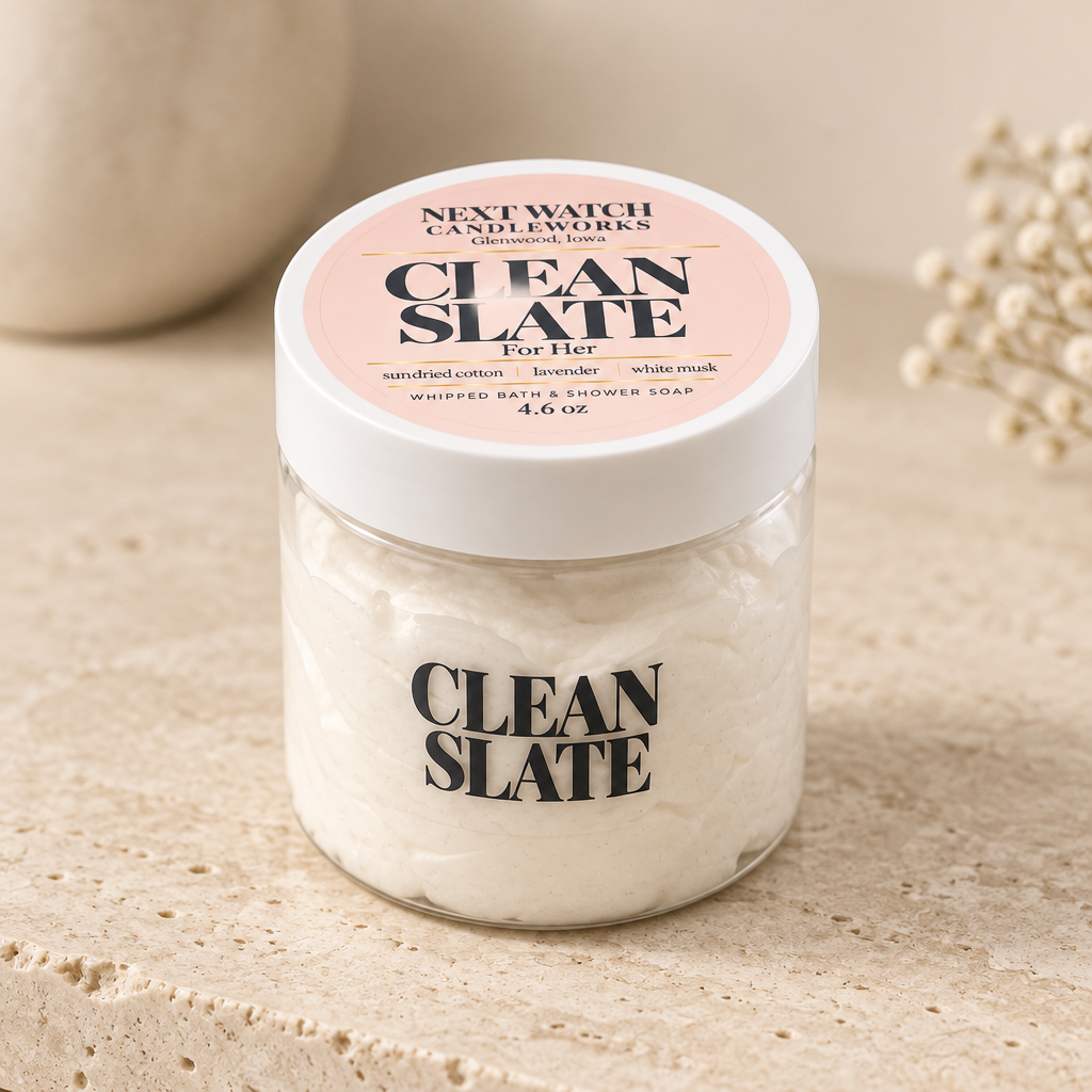 A plastic jar of Next Watch Candleworks Clean Slate For Her whipped bath and shower soap, filled with off-white soap and featuring a pink and white label, set on a textured beige stone surface.