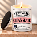 A lit white soy wax candle in a cylindrical jar with a black lid leaning beside it on a stone surface. The front label reads Next Watch Candleworks and Clean Slate, with a pink band and black text.