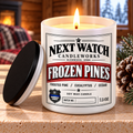 A lit soy wax candle in a white cylindrical jar with a black lid leaning beside it on a stone surface. The front label reads "NEXT WATCH CANDLEWORKS" and "FROZEN PINES," with pinecones and evergreen branches around it and a warm indoor background visible through the window.